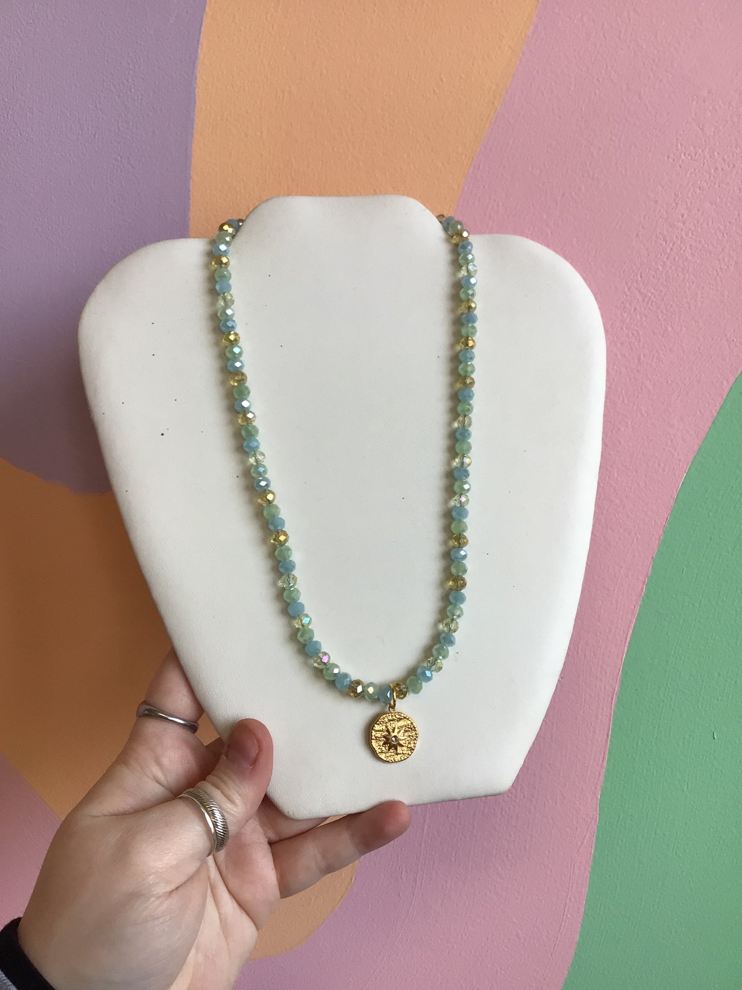A faceted bead glass necklace, with a pattern for clear, gold, pastel blue, and pastel green beads. A textured gold titanium charm of a compass dangles from the center.