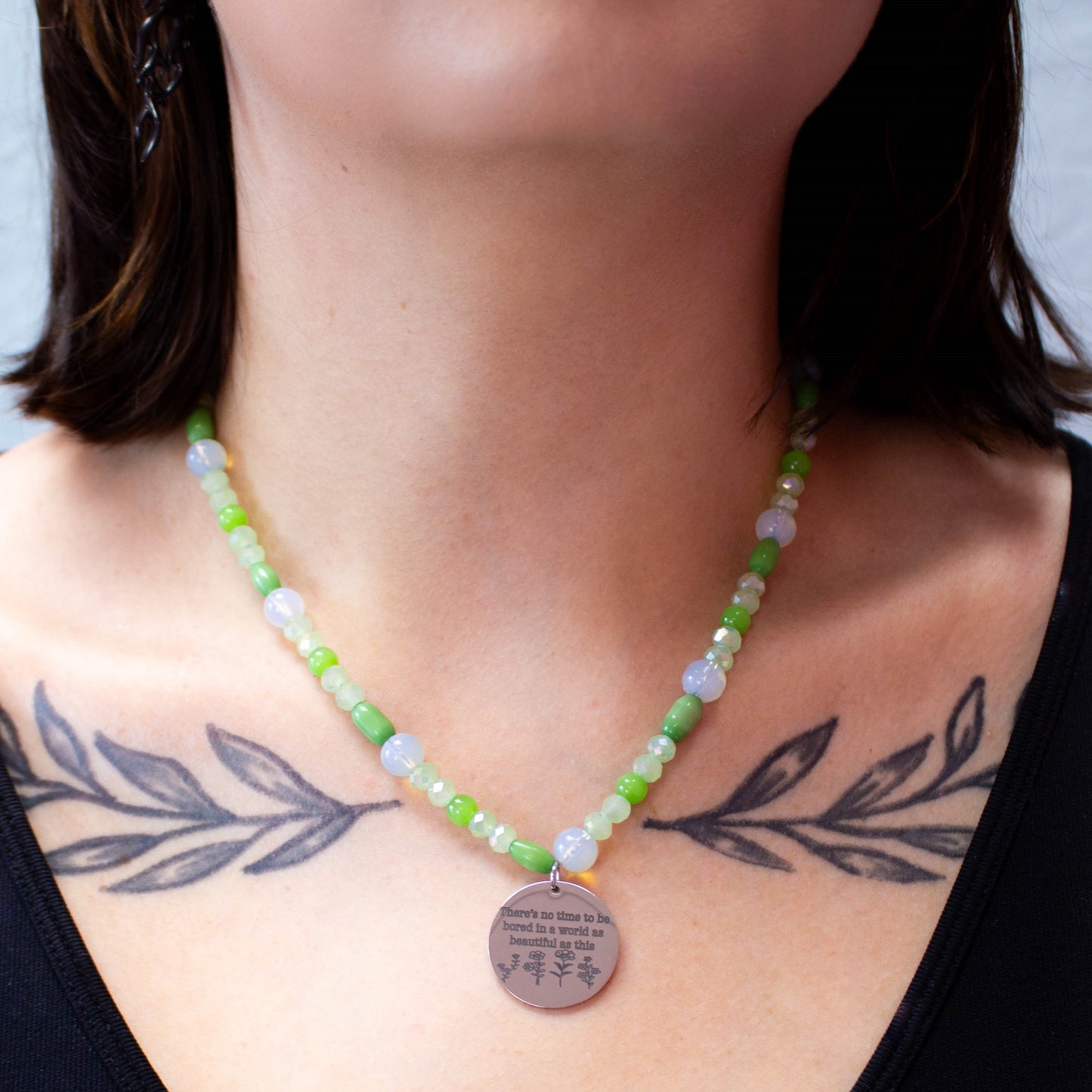 Glass beaded necklace with engraved charm reading "There's no time to be bored in a world as beautiful as this" is shown close up on model's neck