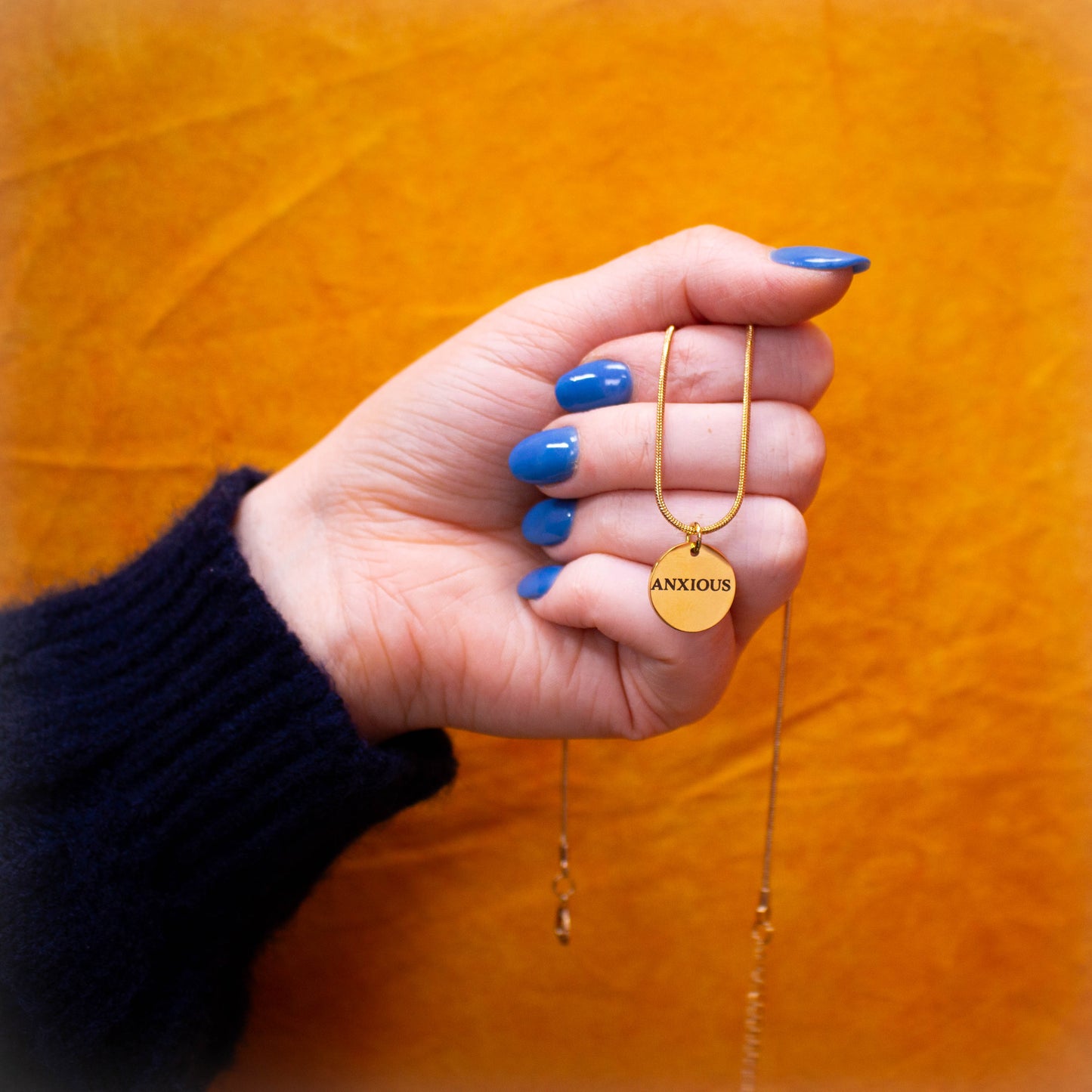 Hand holding engraved "Anxious" necklace in front of mustard yellow backdrop