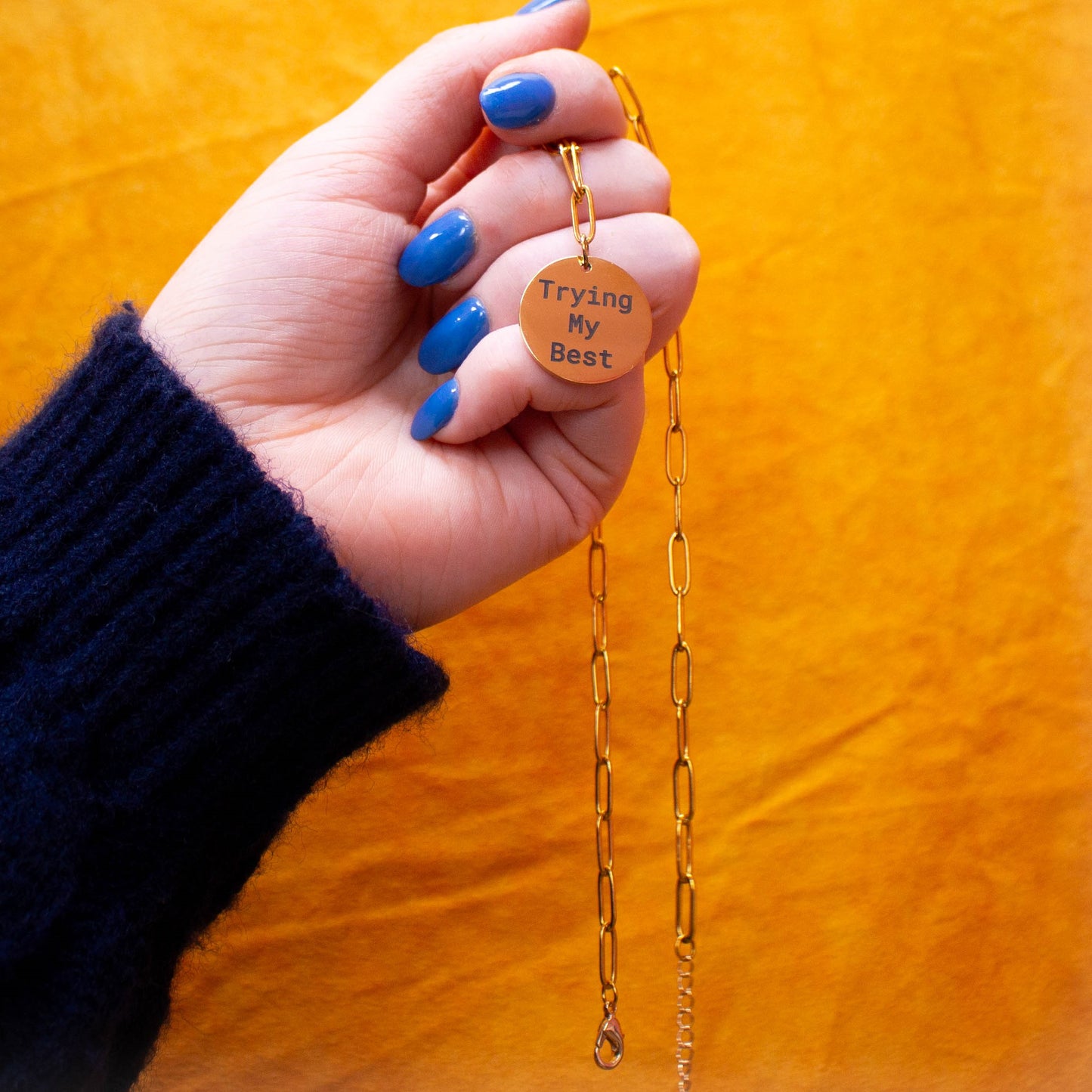 Hand holding "Trying my best" engraved necklace in front of yellow backdrop