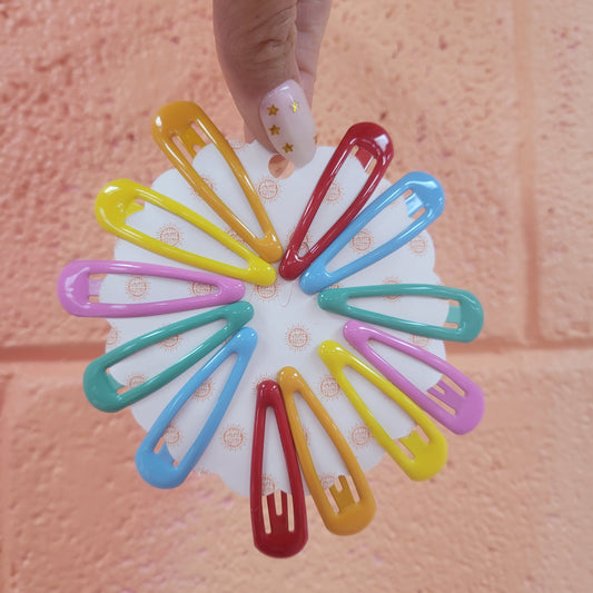A picture showing 12 colorful enamel coated snap clip barrettes. 2 of each in red, orange, yellow, teal, blue, and lilac.