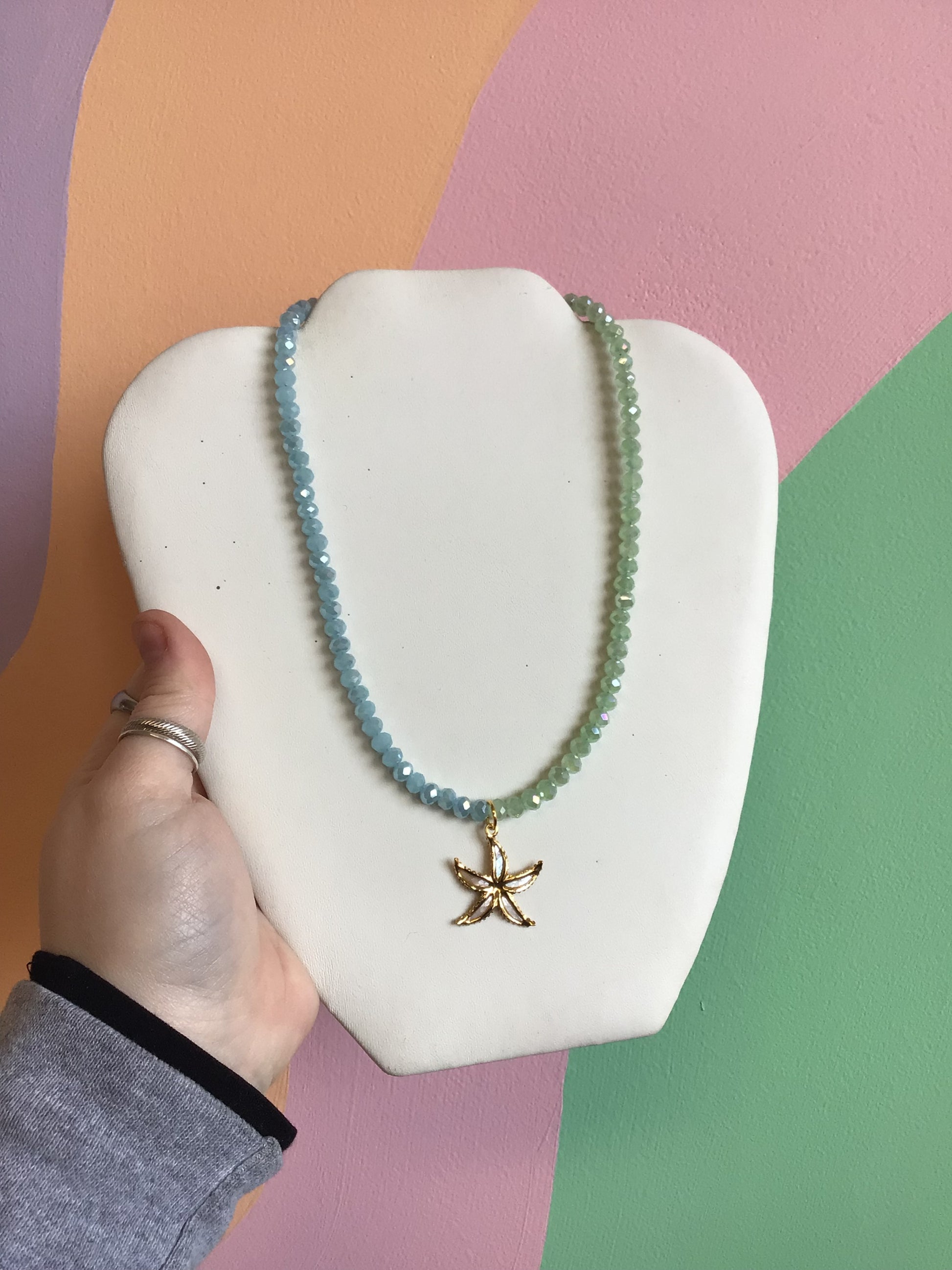 A hand-beaded necklace, where one half is faceted pastel blue beads, the other half pastel green faceted beads. A gold starfish titanium charm dangles from the center, with enamel inlays.