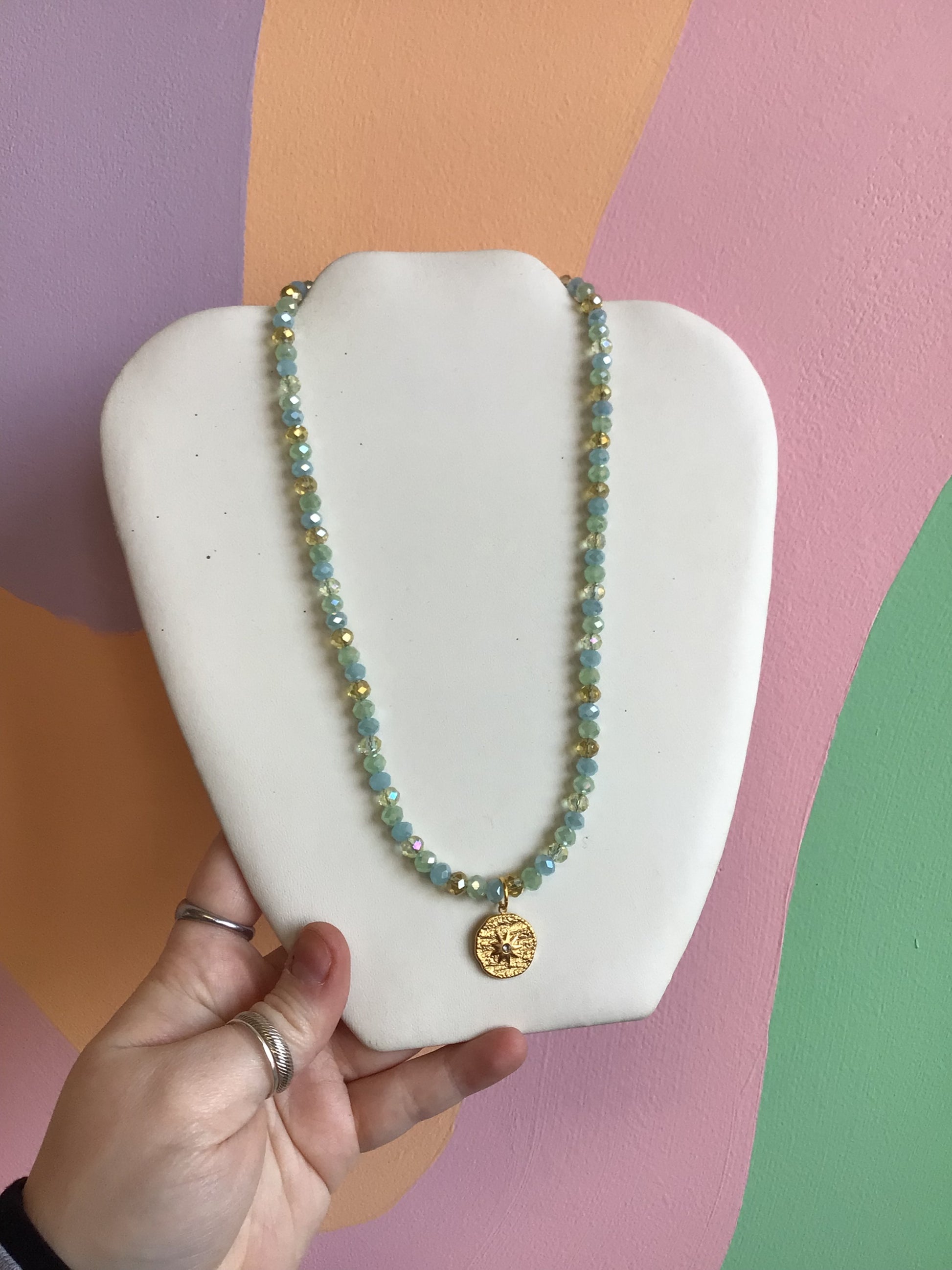 A faceted bead glass necklace, with a pattern for clear, gold, pastel blue, and pastel green beads. A textured gold titanium charm of a compass dangles from the center.