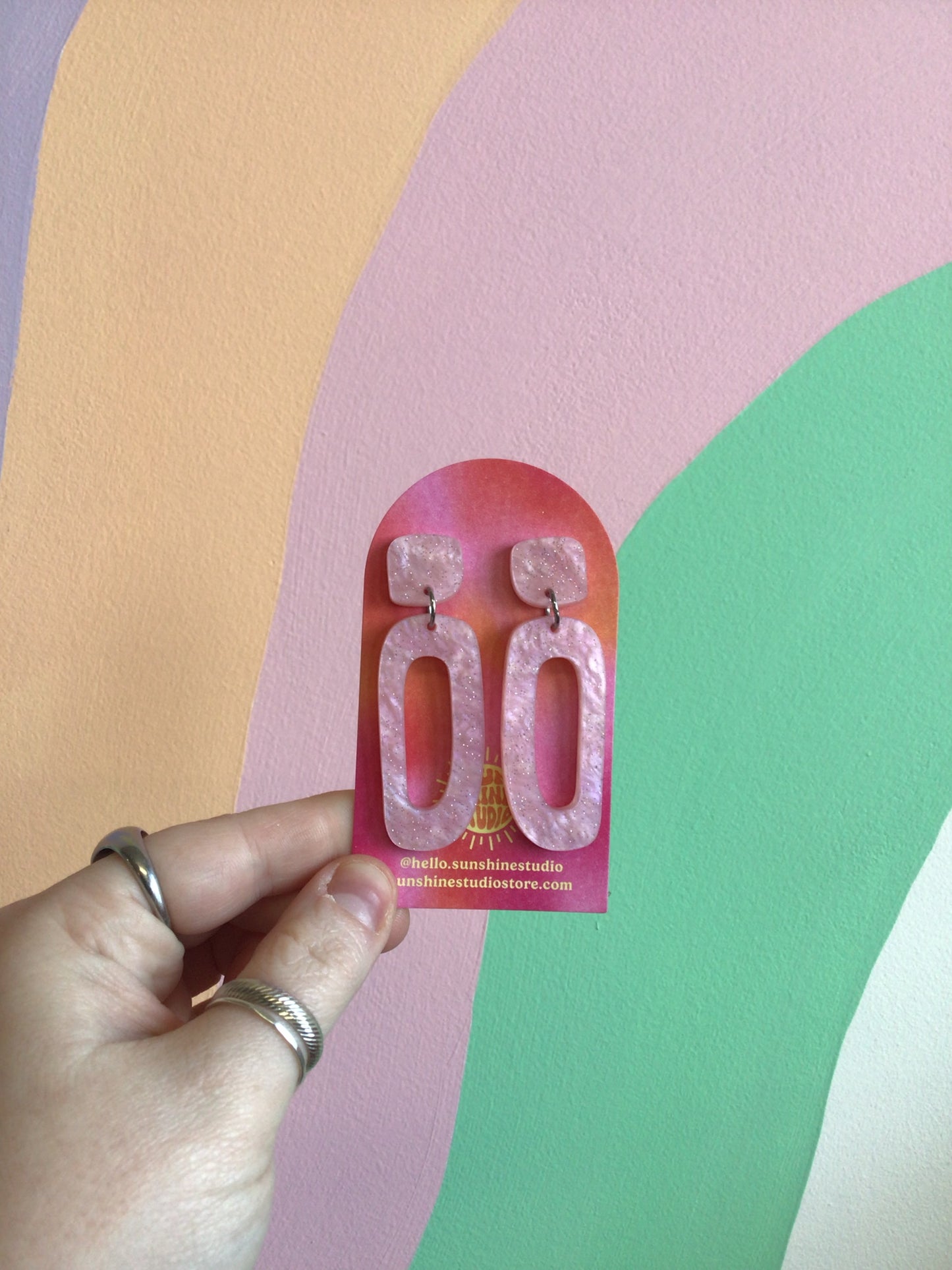 A pair of earrings with an organic cut-out oval shape dangling from a curved square top hiding a stud top. Made of a glittering pink marble acrylic.