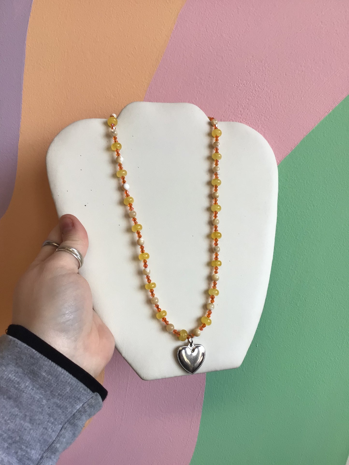 A strung necklace made up of a pattern of yellow and ivory stone beads and small orange glass beads. A silver titanium heart charm hangs from the middle.