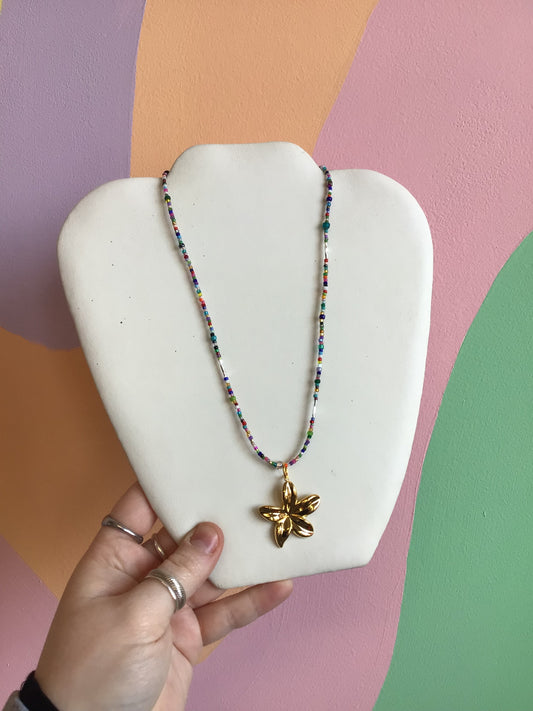 A strung necklace made up of rainbow seed beads and a dangling gold flower titanium charm hanging from the center.