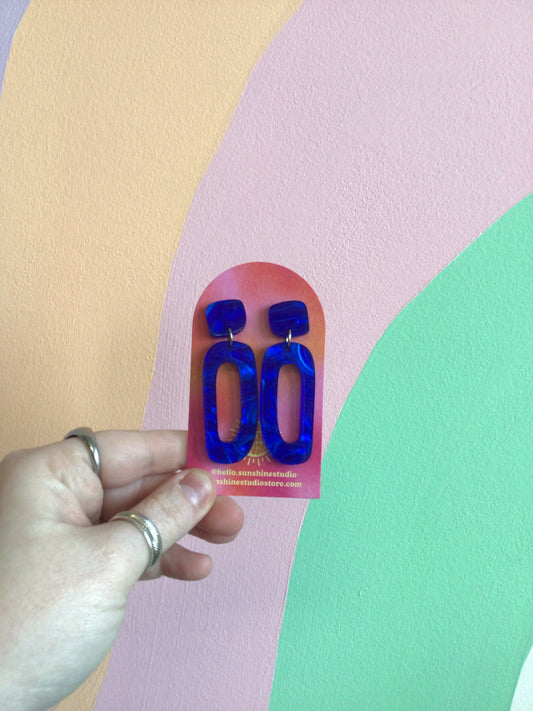 A pair of earrings with an organic cut-out oval shape dangling from a curved square top hiding a stud top. Made of a bright blue pearl finish acrylic.